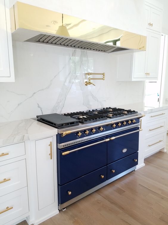 a white kitchen is spruced up with marble, a brass hood and a large cobalt blue cooker