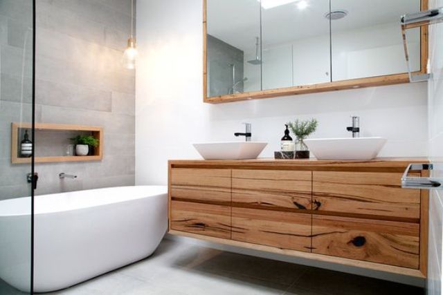 a modern space with a grey tile wall, a wooden vanity and white sinks and a bathtub