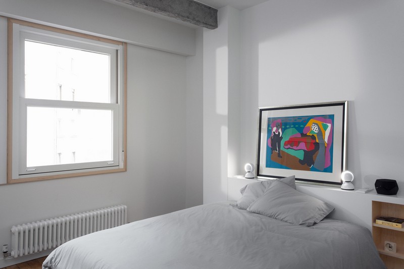 The master bedroom is purely white, there's only a window, a bed, a built in shelf as a headboard and a bold artwork