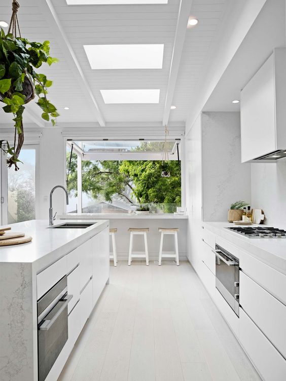 a light white kitchen with marble surfaces and a large window that is extended outdoors as a meal space
