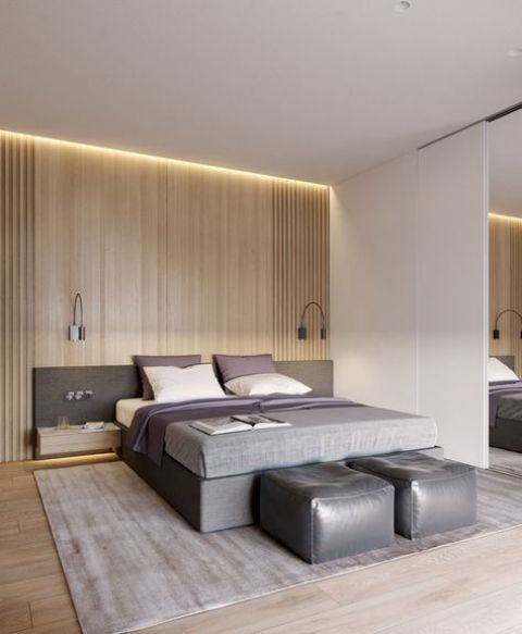 A modern space with light colored wooden wall and floor, an upholstered grey bed with a headboard and grey leather poufs