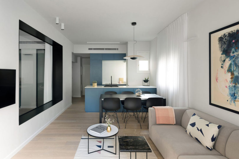 The kitchen and living room are united into one space, the cabinets are done in smoky blue, there's a small dining space with a black dining set and some grey upholstered furniture in the living zone