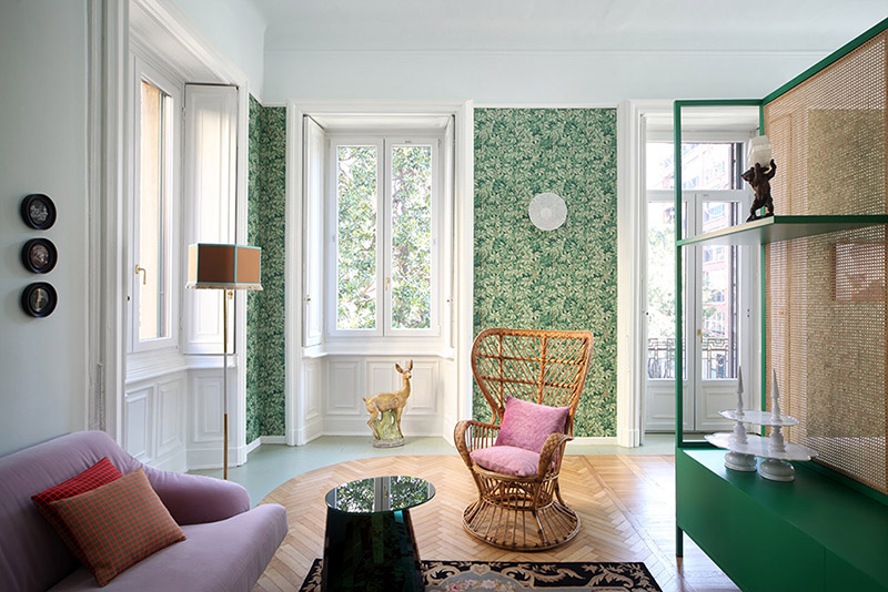 The living room is decorated with botanical print wallpaper, colorful upholstered furniture and a green dresser with a space divider