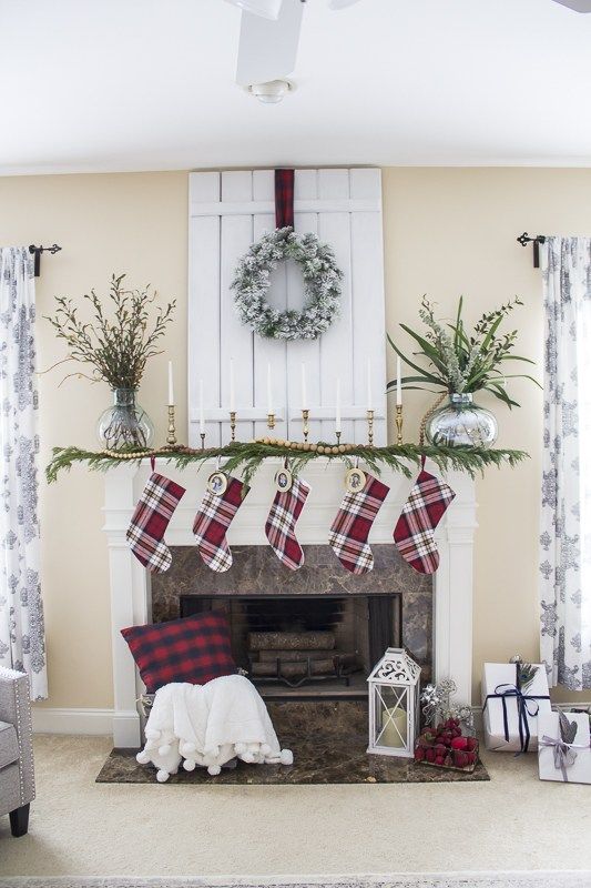 plaid stockings, an evergreen garland with wooden beads, candles in gilded candle holders and a snowy wreath over the mantel
