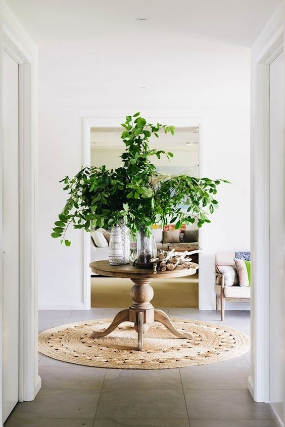 a round pedestal table in the center of the room with greenery in a vase and some wood works as a display table
