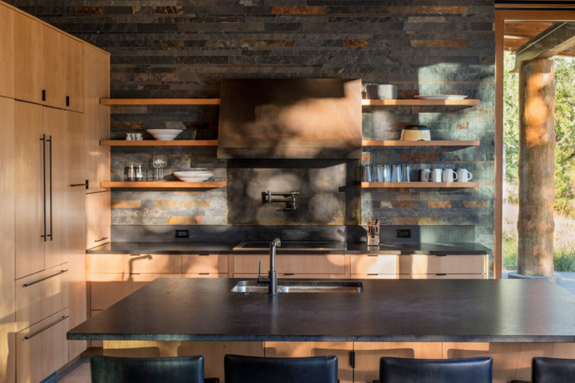 The kitchen is done with light colored wooden cabinets, darkened metal appliances and stone countertops, the walls are clad with reclaimed wood