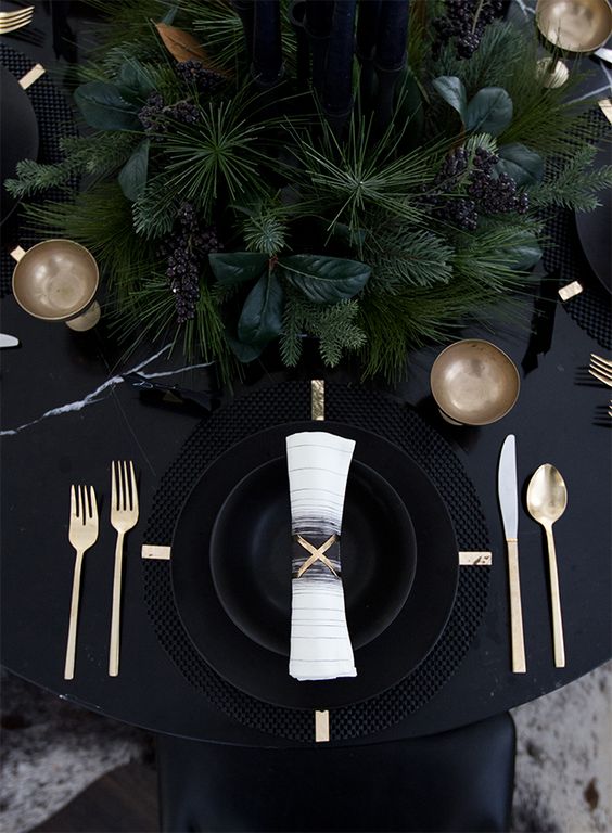 a black table with black plates and cahrgers, lsuh evergreens and gold cutlery for a moody tablescape