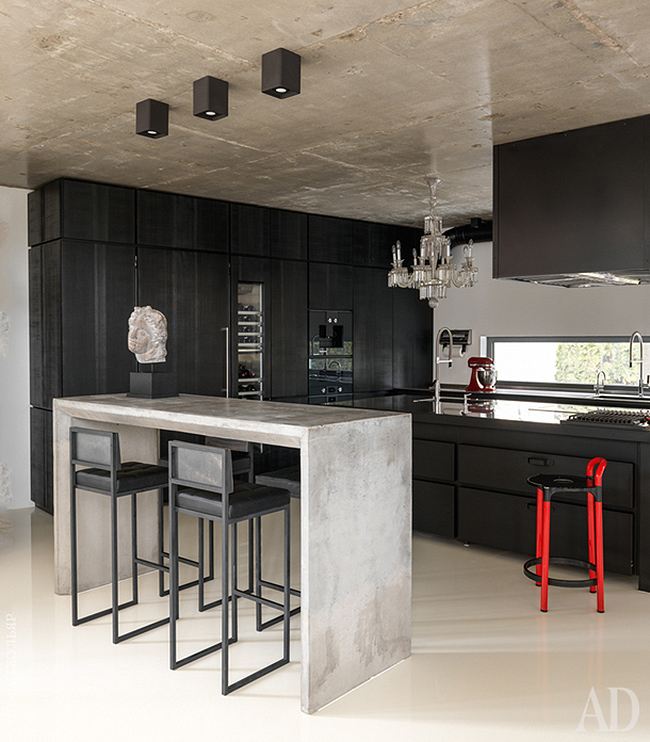 The kitchen is black, in wood and metal, with a crystal chandelier, a concrete console table and some stools