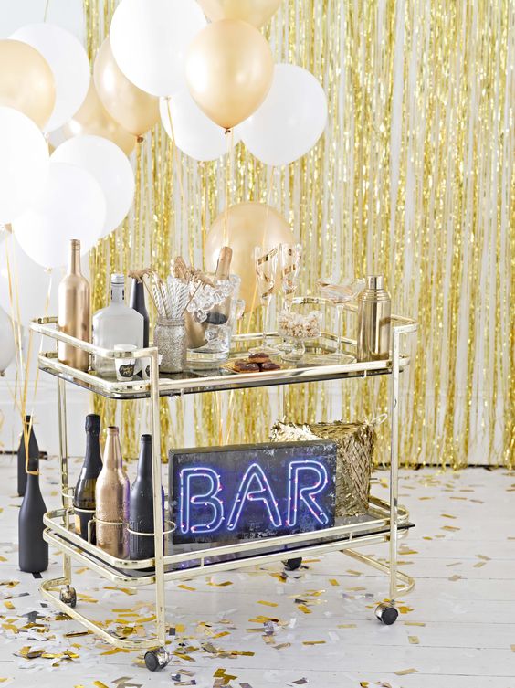 a brass bar cart with white and gold balloons, black and gold bottles and a neon sign
