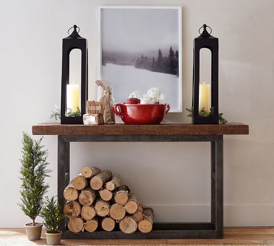tall candle lanterns, a red bowl with ornaments, firewood and evergreen trees under the console