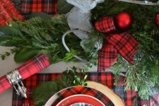 a bright Christmas tablescape with red plaid linens, chargers, ribbon, evergreens and red Christmas ornaments is cheerful