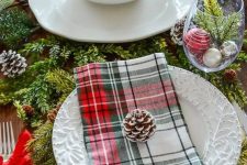 a cozy traditional table setting with evergreens, pinecones, plaid napkins, a tree slice as a placemat and ornaments