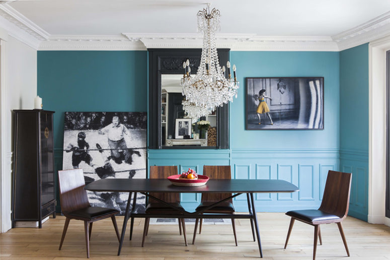 The dining room is done with blue walls, a mid century modern dining set, a glam crystal chandelier and amazing artworks