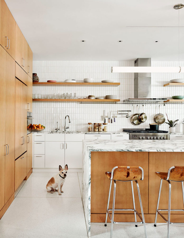 The kitchen united with the dining space is filled with light, done with white tiles and cabinets and some marble