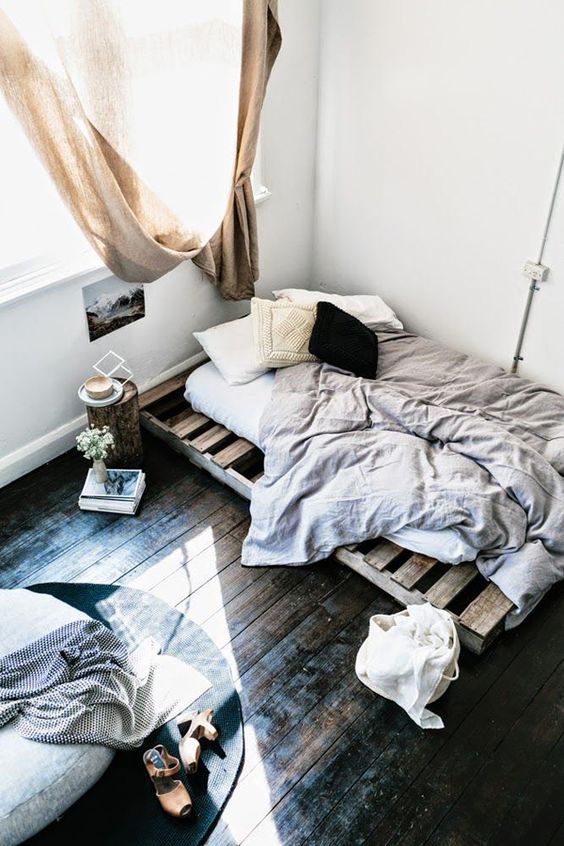 a small industrial bedroom with a pallet bed in the corner and a round ottoman with a rug