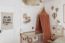 a boho bedroom with a bed and neutral bedding, a crib in the corner and a rust canopy, some decor and toys