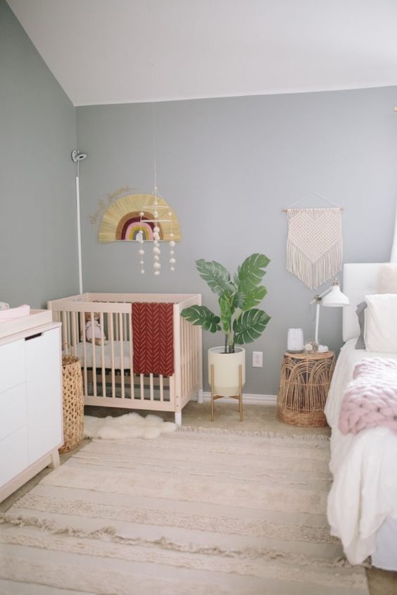 a boho bedroom with grey walls, a white bed and bedding, a crib in the corner, bright bedding, macrame, a potted plant and a dresser