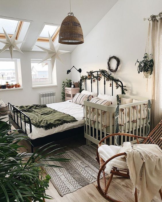 a boho bedroom with skylights, a forged bed with pretty bedding, a pink nightstand, a grey crib, a rocker chair and a pendant lamp