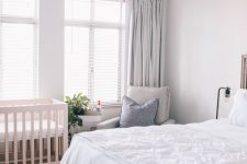 a light-filled bedroom with a bed and white bedding, a crib by the window, a chair and a printed rug