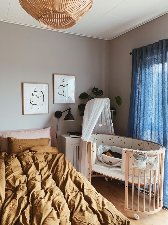 a lovely modern bedroom with grey walls, a bed with mustard and pink bedding, a crib, a nightstand, a wooden pendant lamp and blue curtains