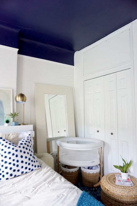 a modern bedroom with a navy ceiling, a bed with a headboard, blue and white bedding, a crib and some baskets