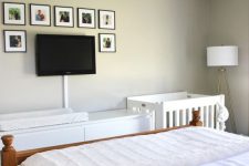 a modern bedroom with a white dresser, a white crib, a floor lamp, a bed with neutral bedding, a gallery wall, a TV
