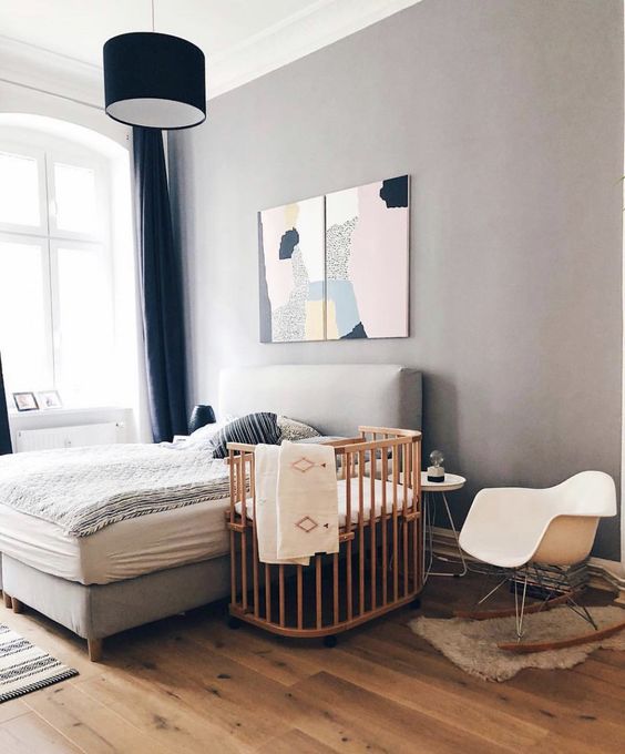 a modern bedroom with grey walls, a grey bed with printed bedding, a crib, a rocker and some art on the wall