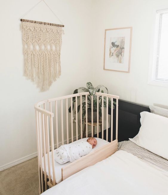 a modern boho bedroom with a bed and neutral bedding, a wooden crib, a stool with a potted plant and macrame
