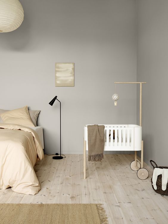 a modern neutral bedroom with a bed and neutral bedding, a crib with a mobile, a black floor lamp and some decor