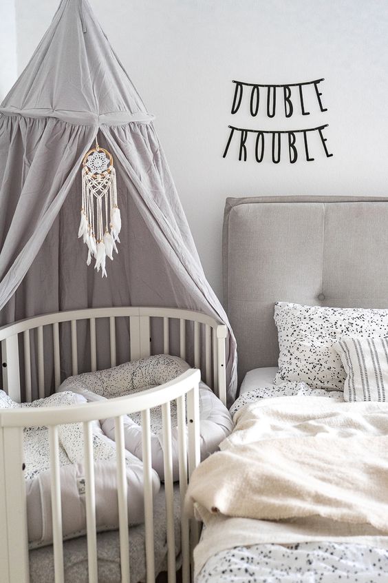 a modern neutral bedroom with a grey bed and printed bedding, an oval crib with a canopy and macrame and some decor