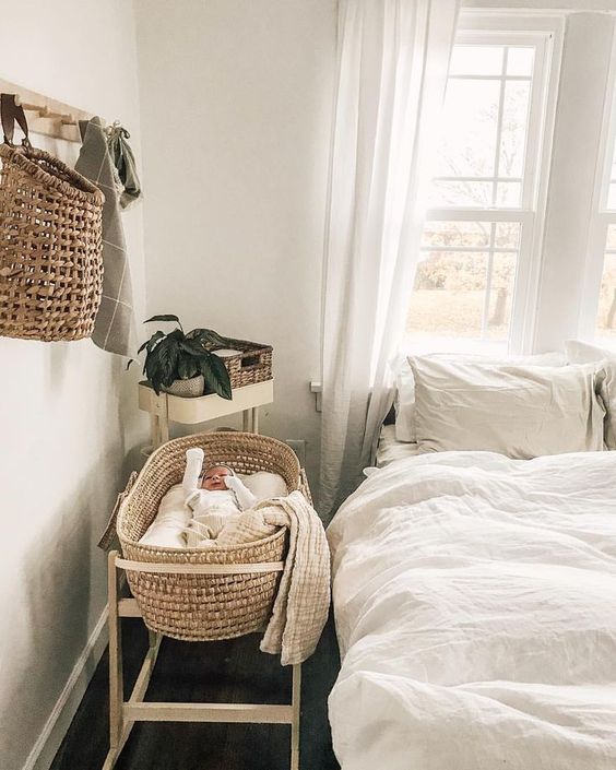 a neutral bedroom with a bed and neutral bedding, a woven crib on a stand, some decor and some greenery