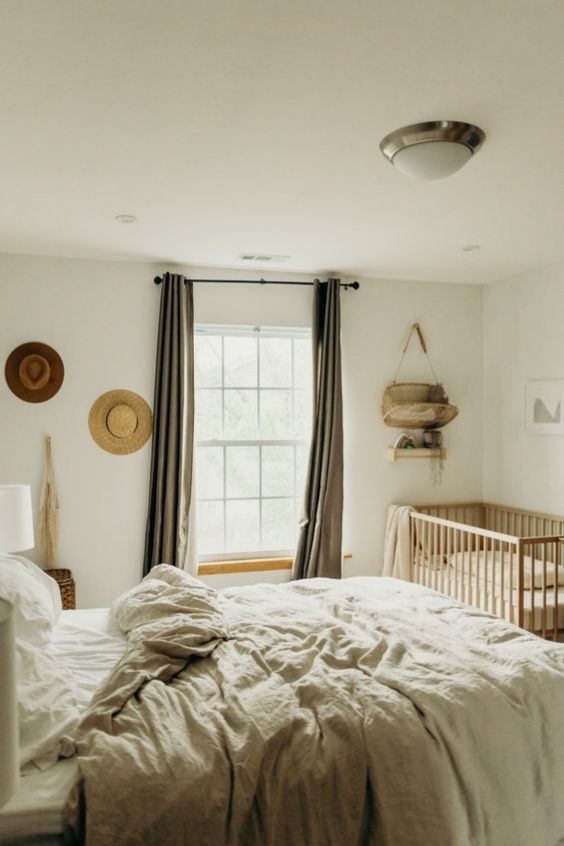 a neutral boho bedroom with a bed and neutral bedding, a crib in the corner, some hats on the wall and some art