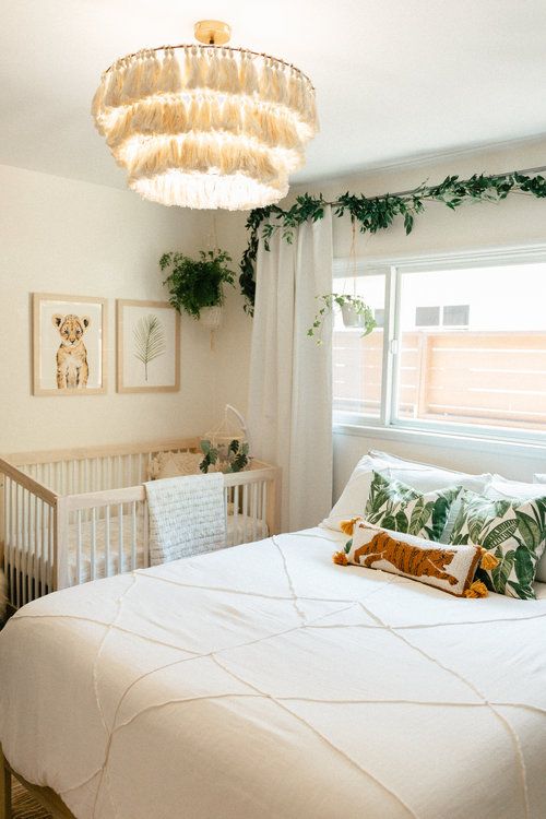 a tropical bedroom with a crib in the corner, a bed with neutral and printed bedding, a tassel chandelier and greenery