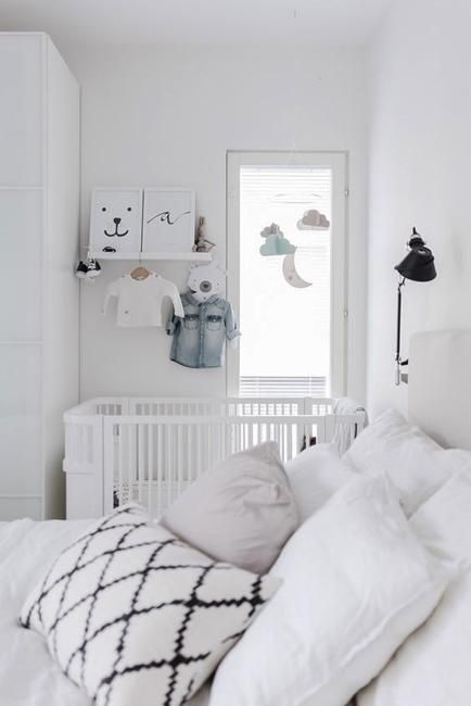 a white Scandinavian bedroom with white furniture, printed bedding, a crib in the corner, a mobile and some decor