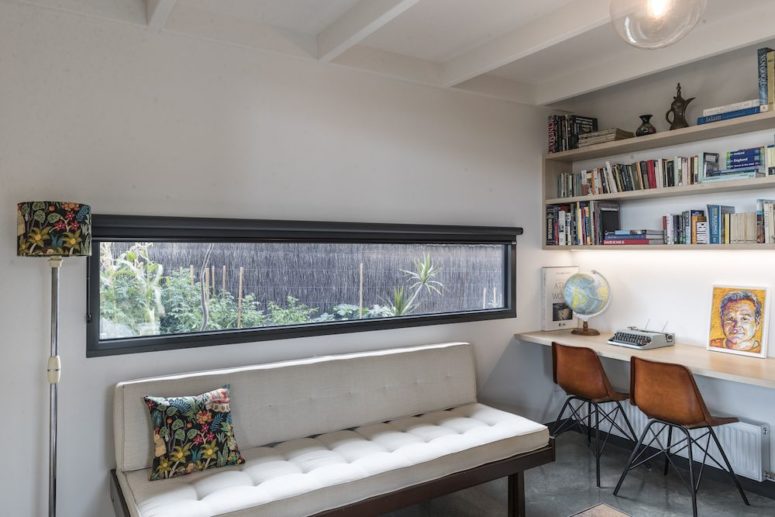 The home office is done with a long narrow window, a shared office space and an upholstered bench