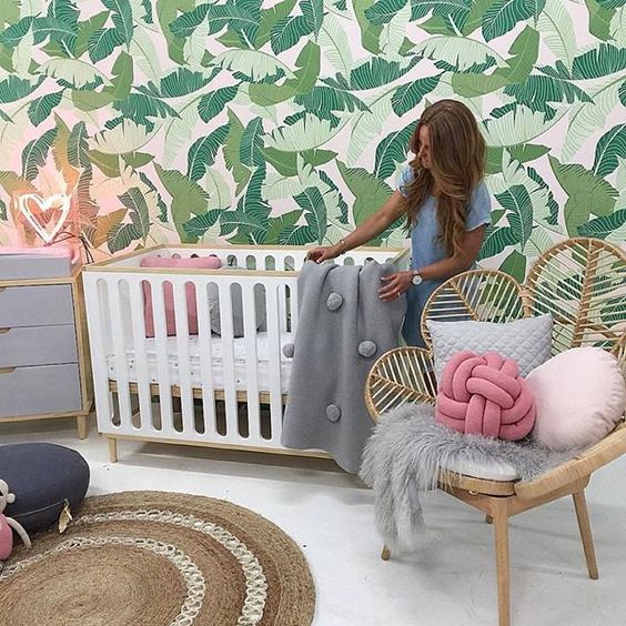 a fun tropical nursery with a tropical leaf statement wall, a rattan chair, a crib, a jute rug and a neon light
