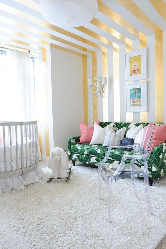 a bright nursery with a glam feel, a striped gold and white wall and ceiling, a tropical leaf print sofa and an acrylic chair