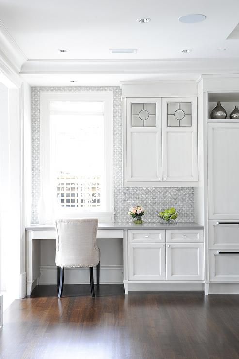 A serene white kitchen with a built in office nook with printed wallpaper to highlight it and a comfy upholstered chair