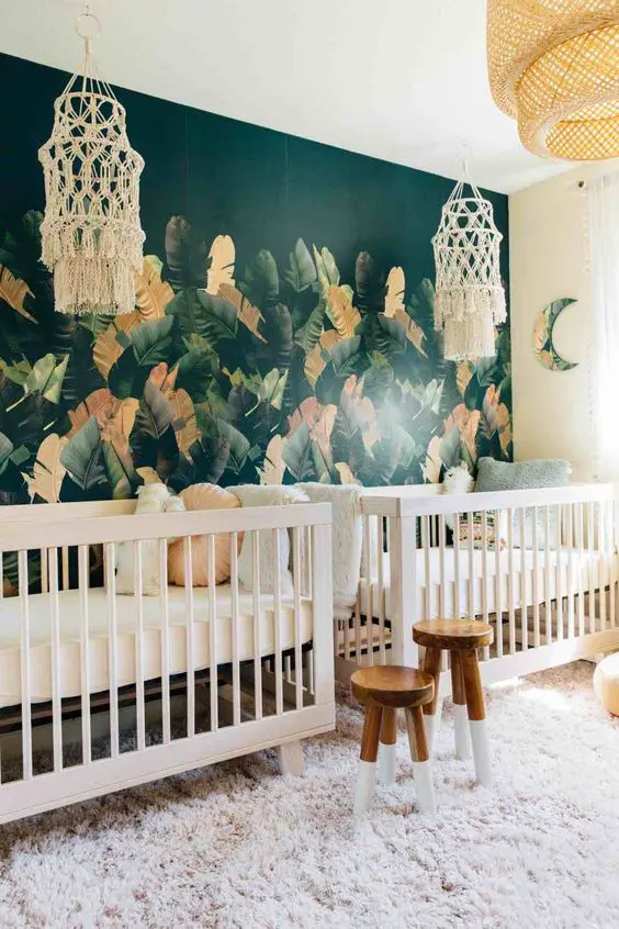 a shared boho tropical nursery with macrame chandeliers, cribs, stools and a faux fur rug plus a statement torpical leaf wall