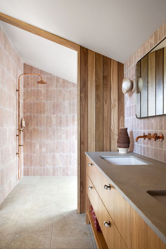 a blush bathroom clad with stone and skinny pink tiles, a double vanity, a wooden divider and some mirrors is cool