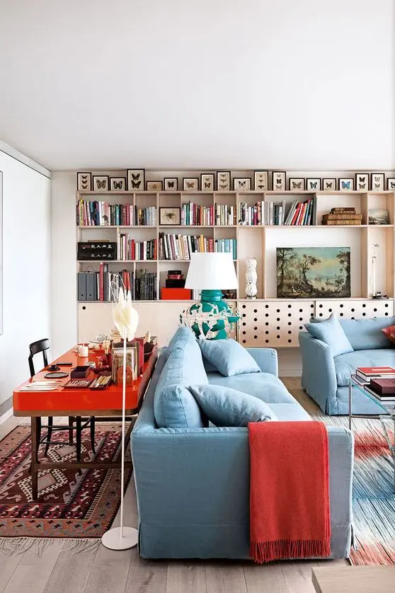 a bold living room with a plywood storage unit on the wall, butterflies, blue furniture, a hot red desk behind the sofa and bold red textiles