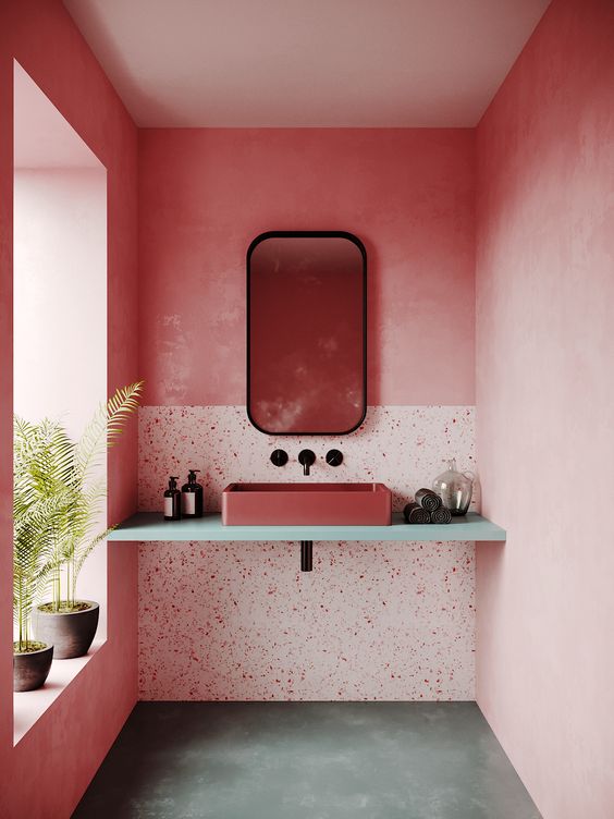 a bright pink bathroom with a terrazzo accent, a terracotta sink on the vanity, potted greenery and a mirror in a black frame
