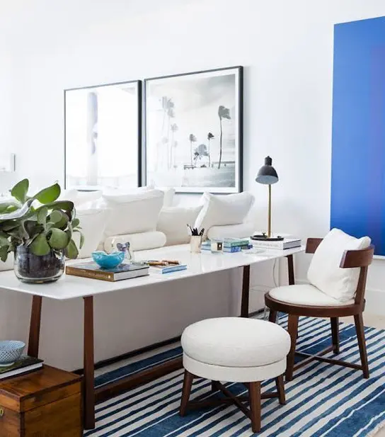 a coastal living room with a bold blue accent wall, creamy furniture, a desk and a chair with a pouf and blue touches