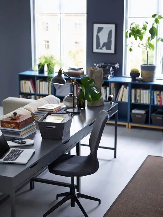 a cool living room with a slate grey wall, blue bookshelves, a neutral sofa, black desk, a black chair, a brown rug and potted plants