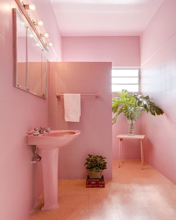 A light pink bathroom clad with square tiles, a free standing sink, a shower space, a mirror with lights and potted greenery