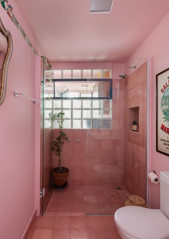 a light pink bathroom with coral tiles in the shower, a window and white appliances and greenery is cool and bright