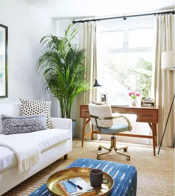 a modern neutral living room with a working space at the window, a creamy sofa, a blue pouf and statement potted plants