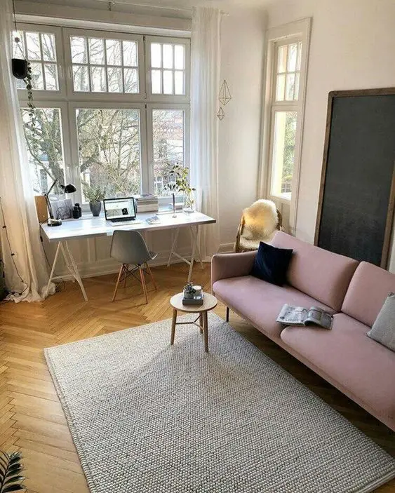 a neutral living room with a trestle desk and a grey chair, a pink sofa and pillows, a side table, a rug is a cozy and lovely space