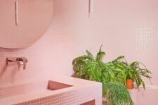 a pink bathroom with small scale tiles all over the space, a round mirror, potted greenery and a vanity clad with tiles, too
