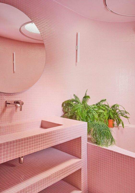 a pink bathroom with small scale tiles all over the space, a round mirror, potted greenery and a vanity clad with tiles, too